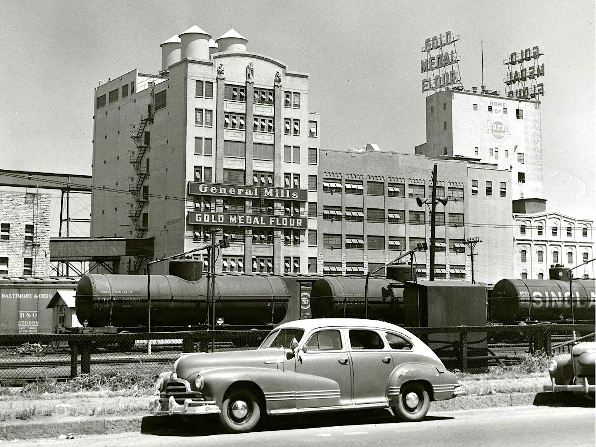 A black and white photograph shows the General Mills building with "General Mills" and "Gold Medal Flour" signs prominently displayed. On the roof of a taller building behind it, large signs spell out "GOLD MEDAL FLOUR." In the foreground, a vintage car is parked on the side of a road, and behind it, several train cars, including tank cars and a boxcar are visible on tracks.